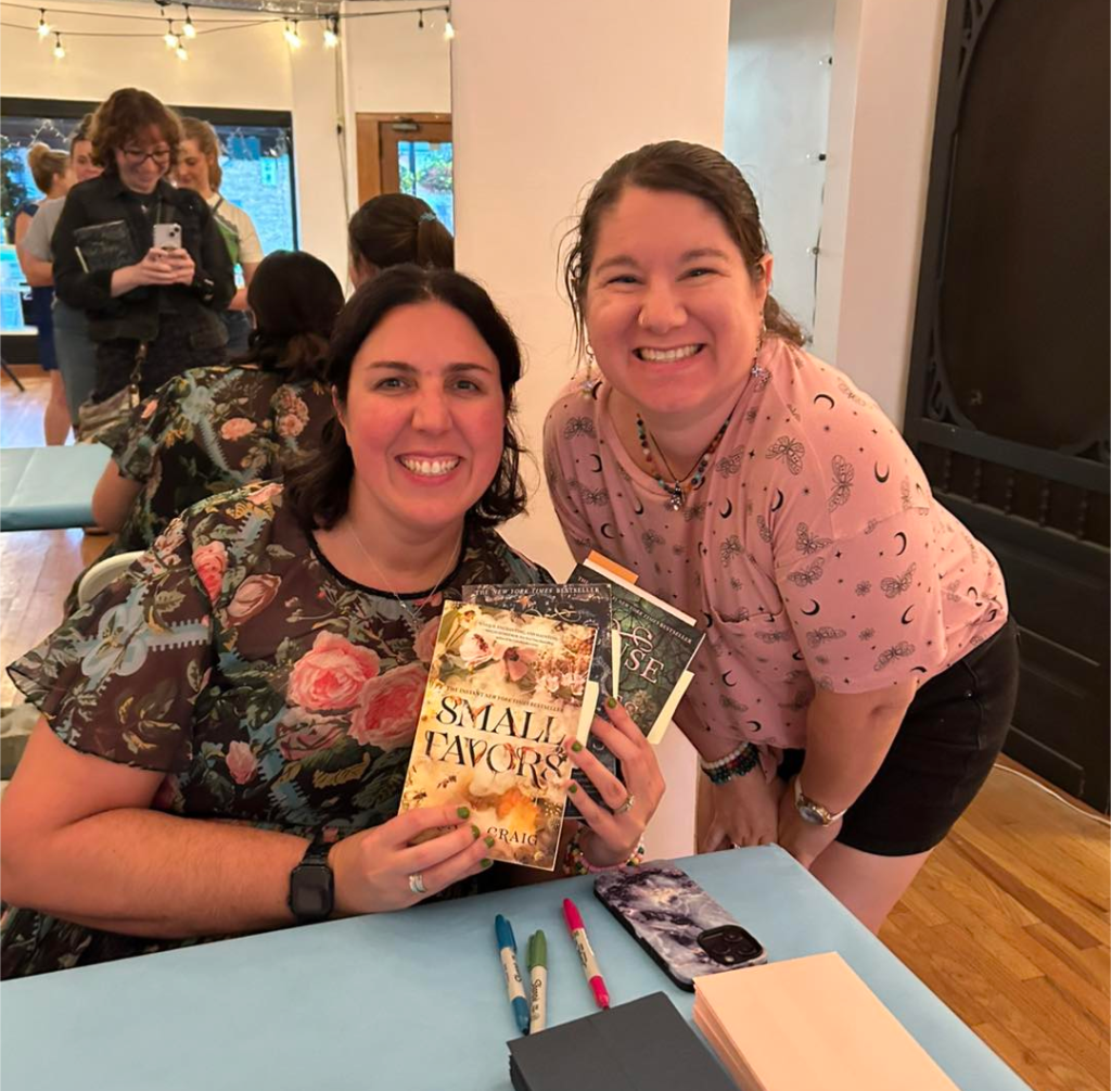 Image of Lena (right) crouching beside author Erin Craig (left) who is seated and holding three books. A mirror behind them shows someone taking the photo and a line of people waiting for their books to be signed.