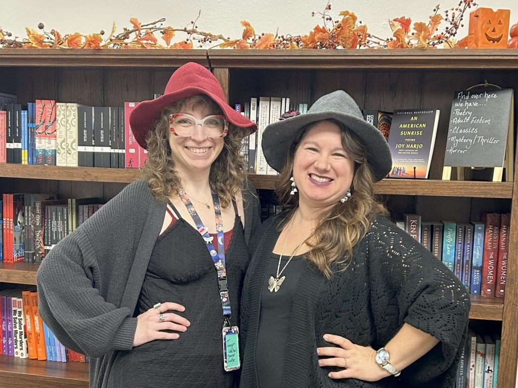 Tracy (left) and Lena (right) dressed as 90s witches for District Frights posing in front of bookshelves decorated for Autumn