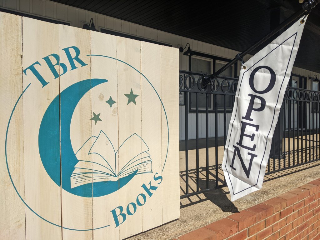 Large wooden sign with the TBR Books Logo in bluegreen with an Open flag beside it