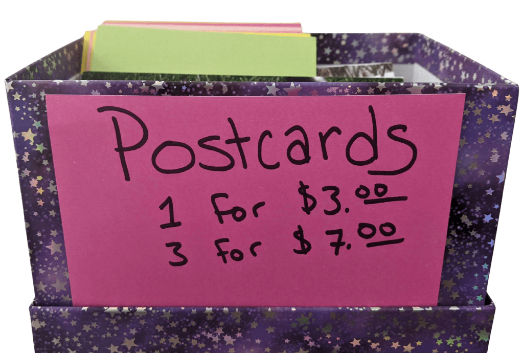 An open box decorated in purple with shiny silver stars, labeled with a hot pink card stating "postcards, 1 for $3.00, 3 for $7.00."