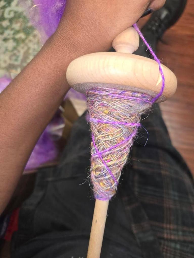 Rownan's hand holding a drop spindle with newly spun yarn on it