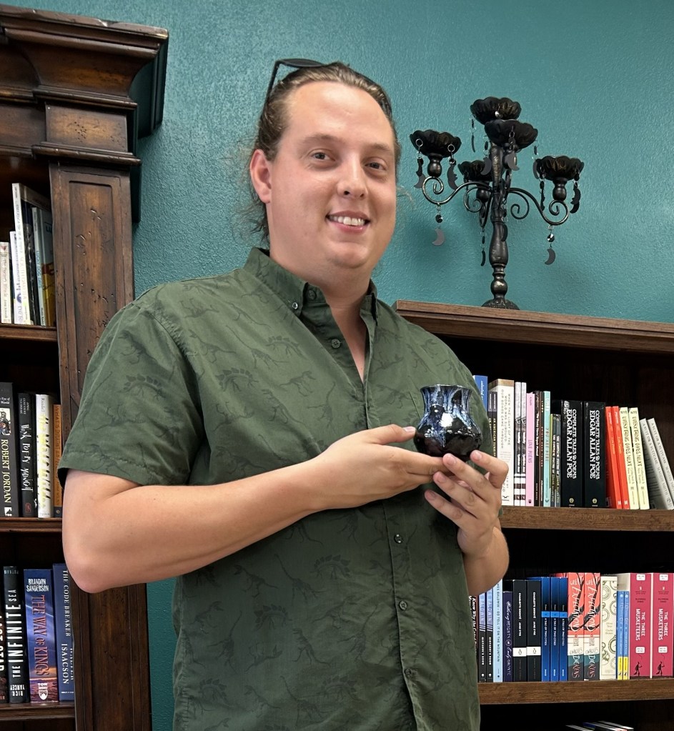 Ceramic Bob holding one of his pottery pieces in front of a bookshelf and candelabra at TBR Books.