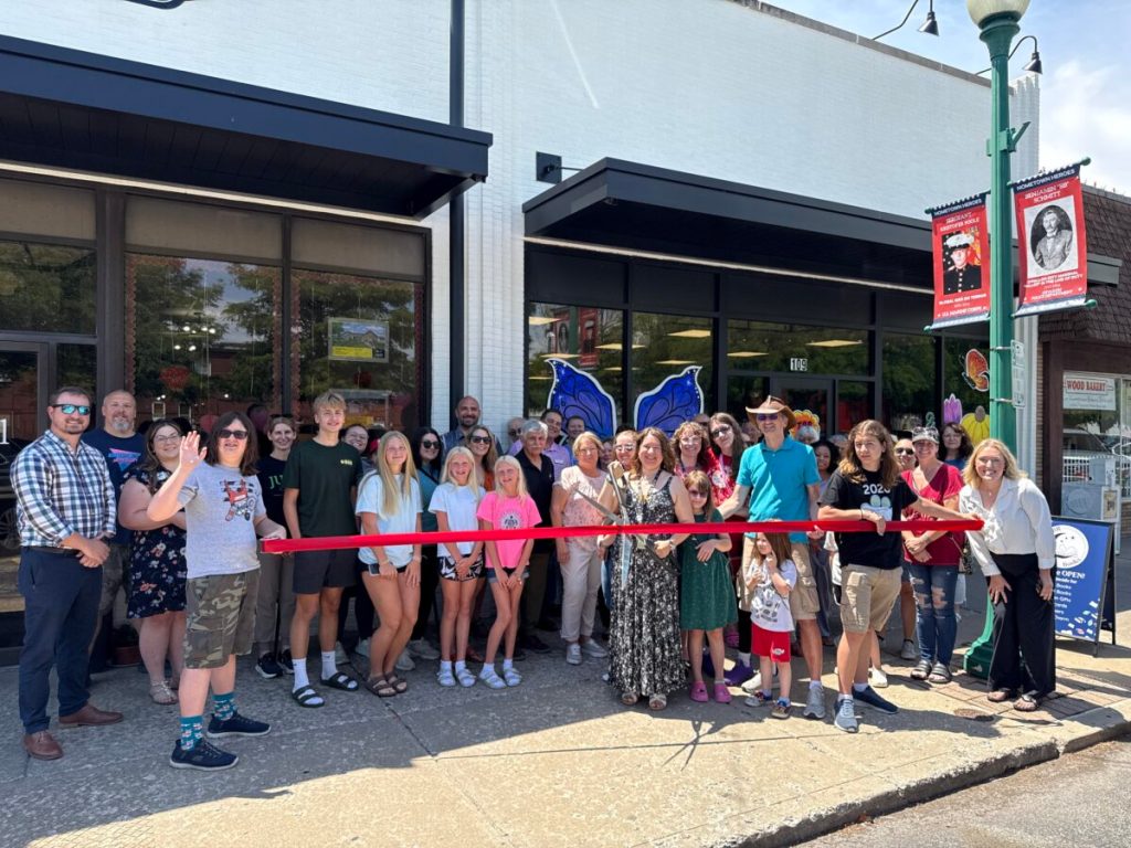 Ribbon cutting ceremony: image of Lena, her family and friends, and supportive supportive customers clustered behind a red ribbon in front of TBR Books storefront. Lena is holding the big scissors.