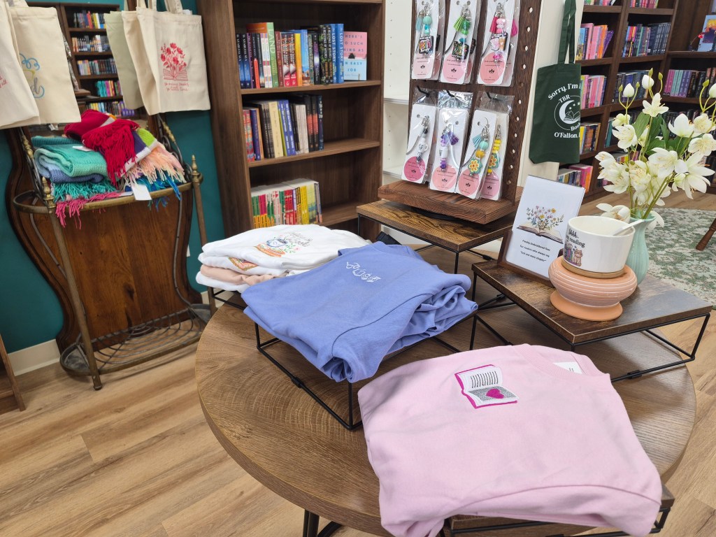Simply Stylish Stitches display showing folded t-shirt stacks, bookmarks, and embroidered bags in the background