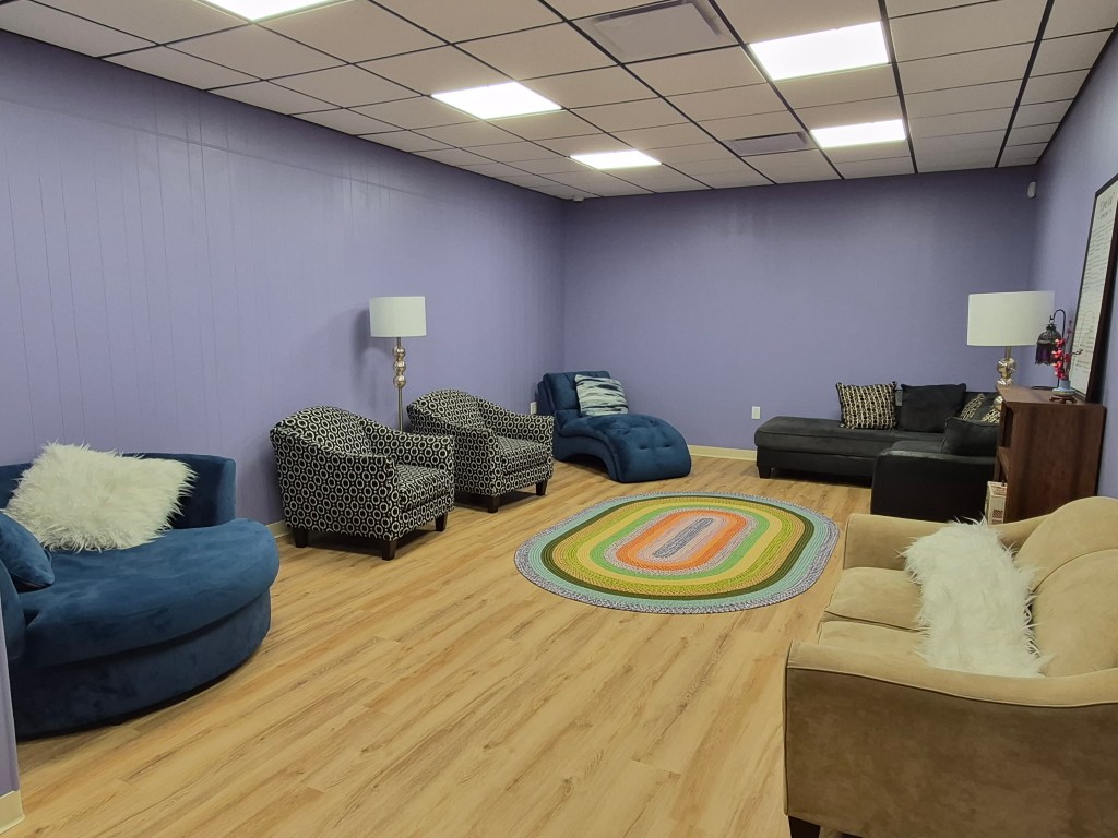 Reading Room at TBR Books: light wood flooring, purple walls, white and black drop ceiling, cozy couches and chairs, and a rug in the middle
