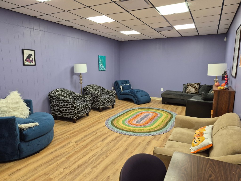 Photo of a room with lavender walls, a light wood floor, and a black and white drop ceiling. There are a variety of comfortable chairs and sofas, lamps, and art on the wall.